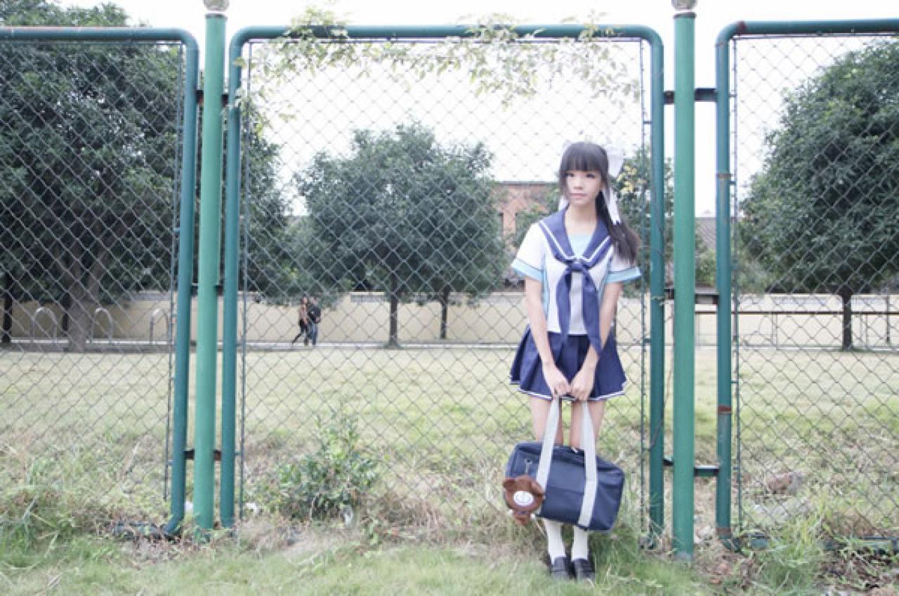 熊酱穿着蓝色学院校服扮演高岭爱花，靠着操场周围的铁丝网围栏站立的样子