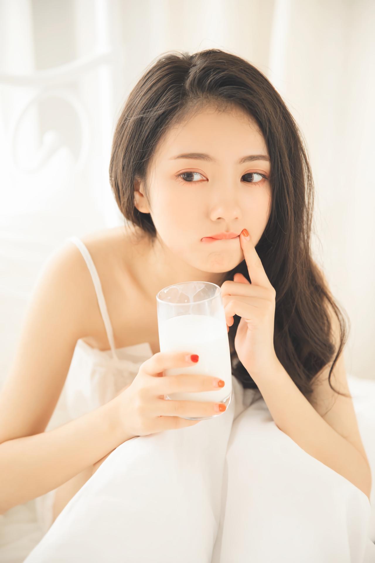 少女坐在床上，右手握着牛奶杯，作手食指抵着唇角