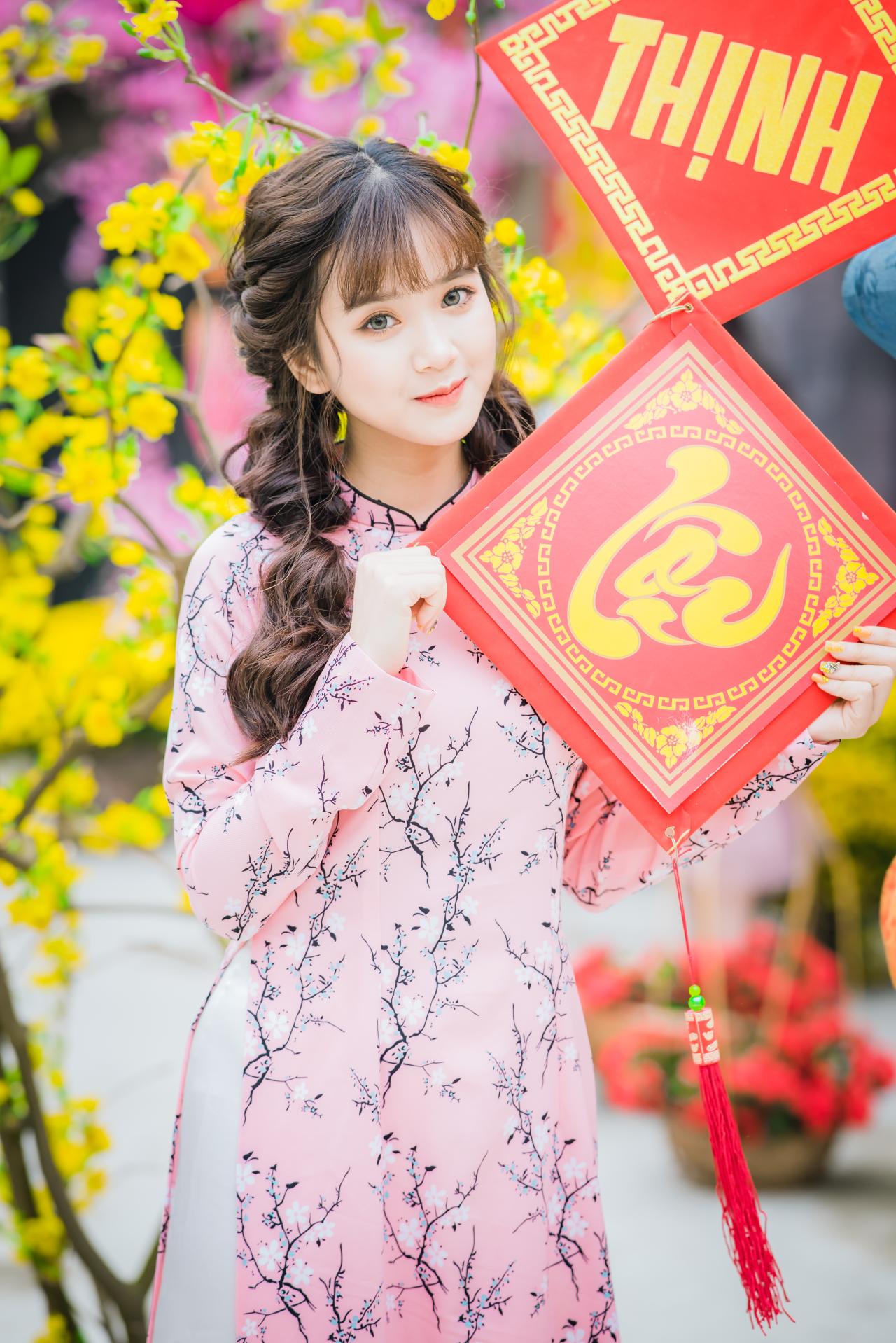 棕色双马尾卷发，齐刘海儿，穿着粉色旗袍的越南少女 站姿 全身照 正面照 竖幅 双手抱着红色福字中国结 微笑 腊梅 黄梅 树