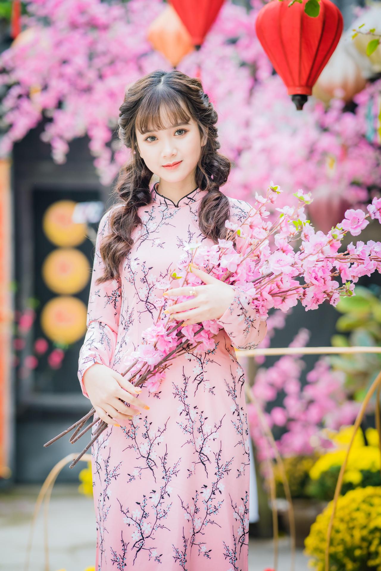 棕色双马尾卷发，齐刘海儿，穿着粉色旗袍的越南少女 站姿 全身照 正面照 竖幅 双手捧着一束粉色樱花 微笑 腊梅 黄梅 花篮 红色灯笼