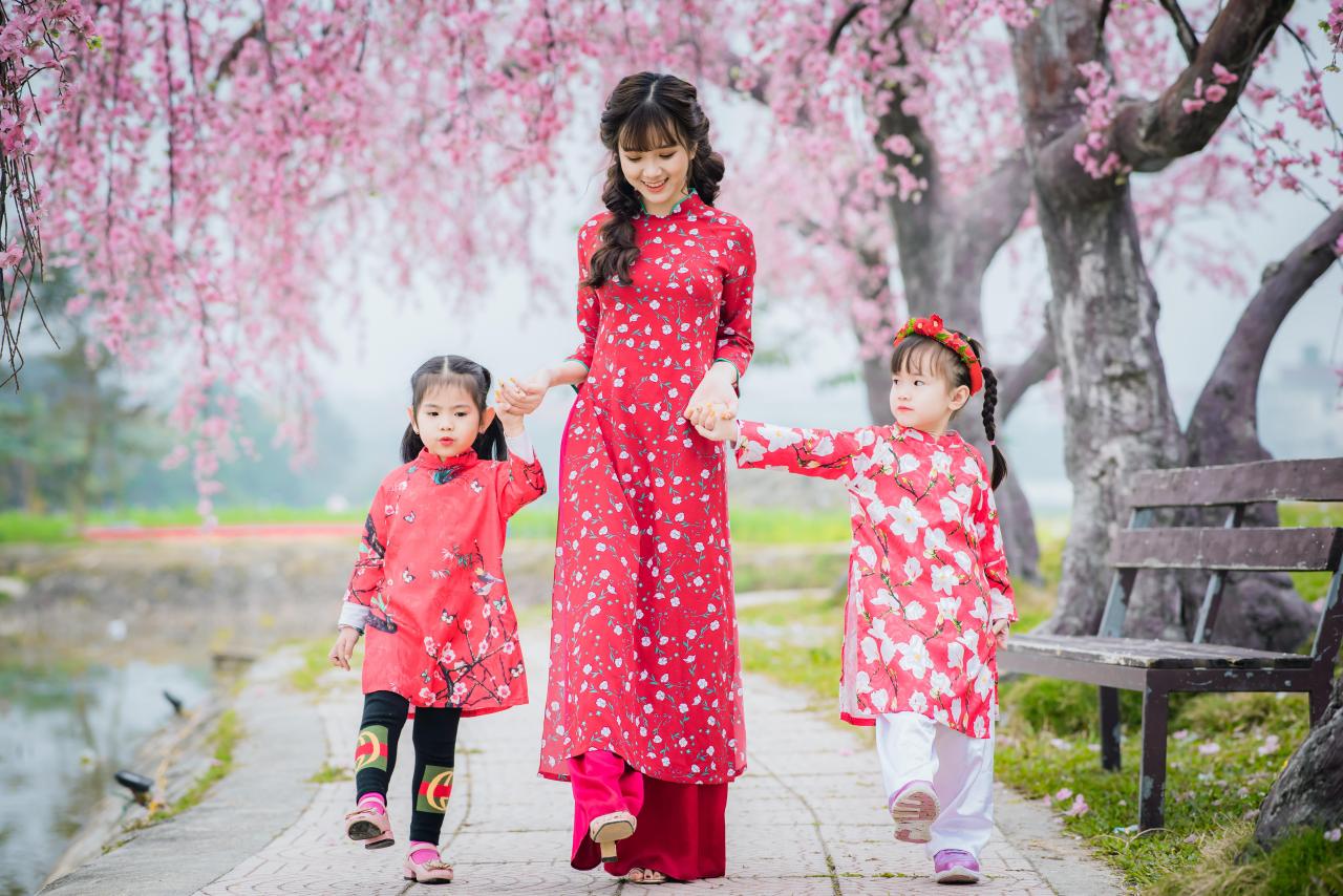 棕色双马尾卷发，齐刘海儿，穿着红色旗袍的越南少女手牵着两个小女孩，走在樱花树下的河边石路上 低头微笑 站姿 全身身照 正面照 横幅 樱花 草地 公园长椅