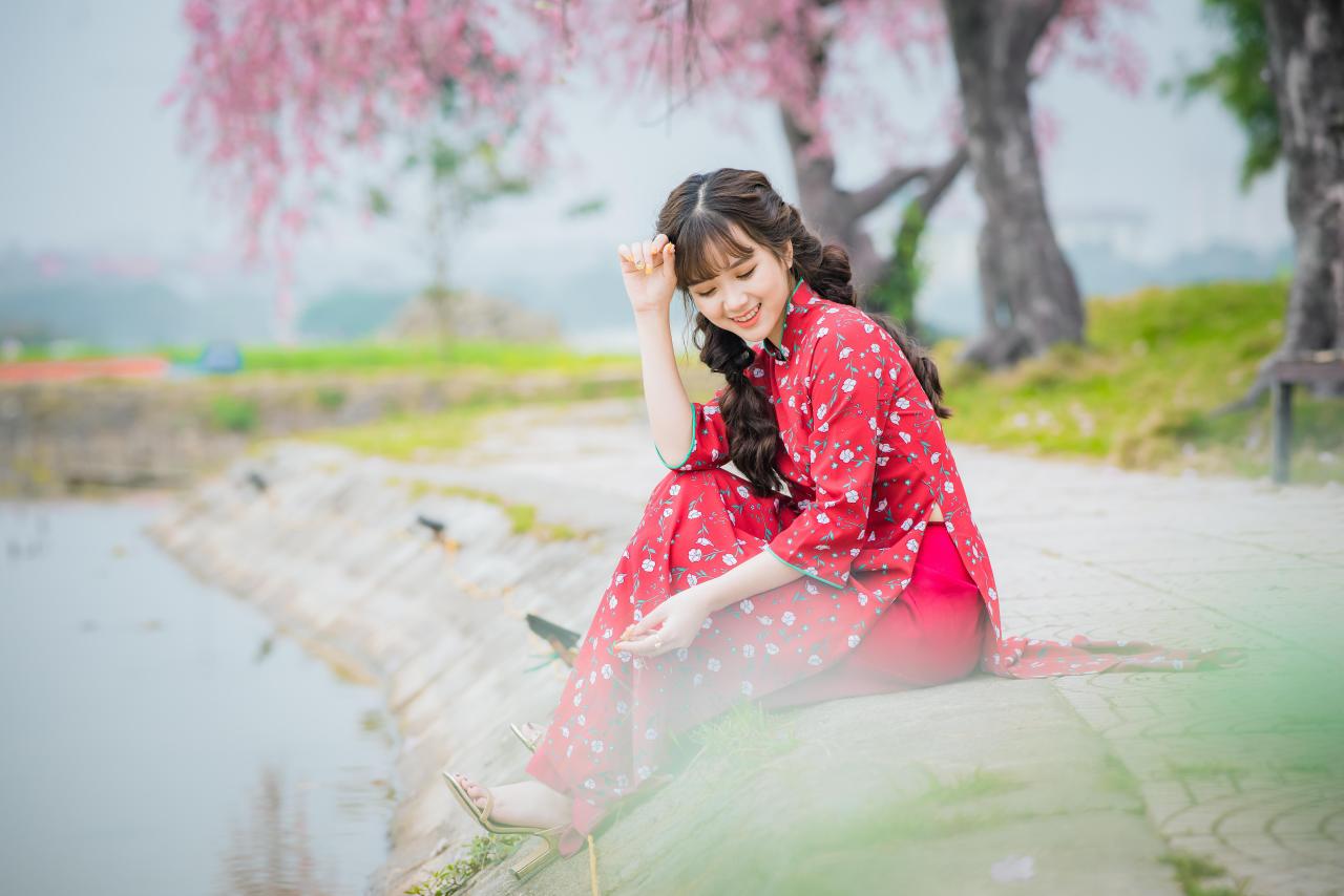 棕色双马尾卷发，齐刘海儿，穿着红色旗袍的越南少女坐在河边 低头咧嘴露齿微笑 轻抚额头 坐姿 全身照 4/3侧面照 横幅 樱花 草地 公园长椅 背景虚化