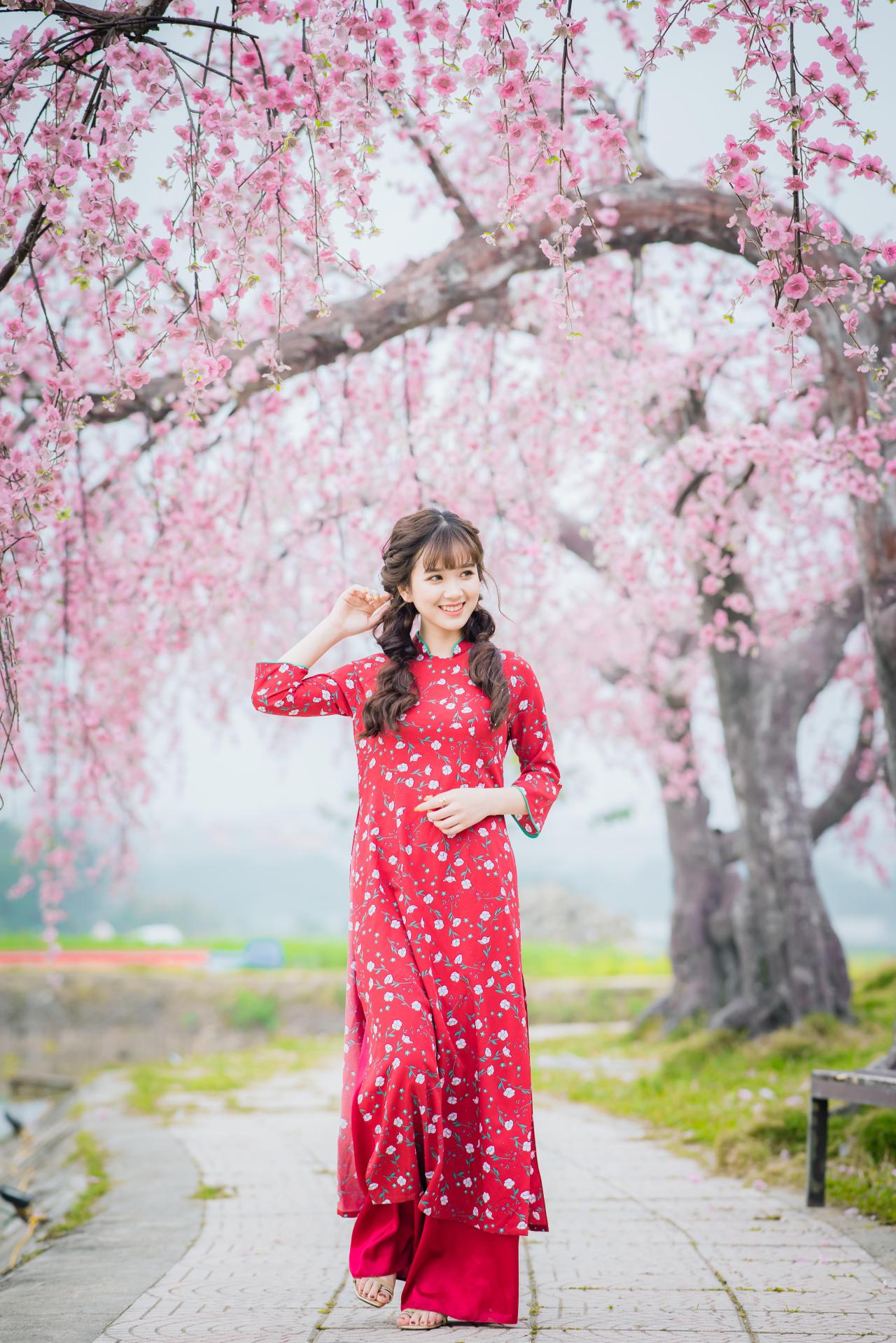 棕色双马尾卷发，齐刘海儿，穿着红色旗袍的越南少女走在樱花树下的石路上 右手轻抚马尾 左手放在腰间 咧嘴露齿微笑 站姿 全身照 正面照 竖幅