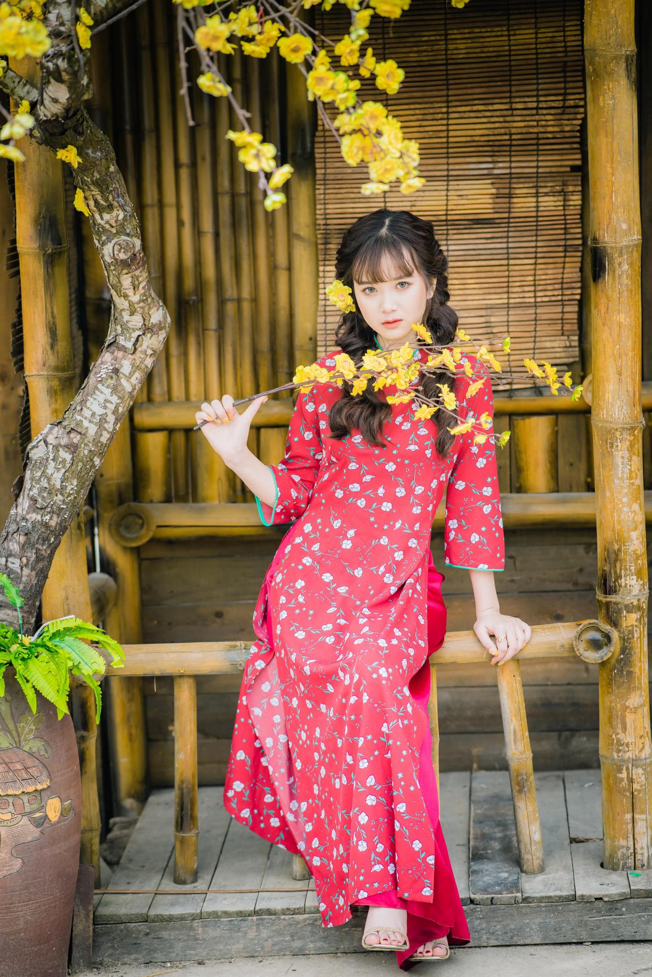 棕色双马尾卷发，齐刘海儿，穿着红色旗袍的越南少女坐在竹屋前 右手拿着一束黄色腊梅花横在胸前 坐姿 全身照 正面照 竖幅 腊梅花树 黄梅树
