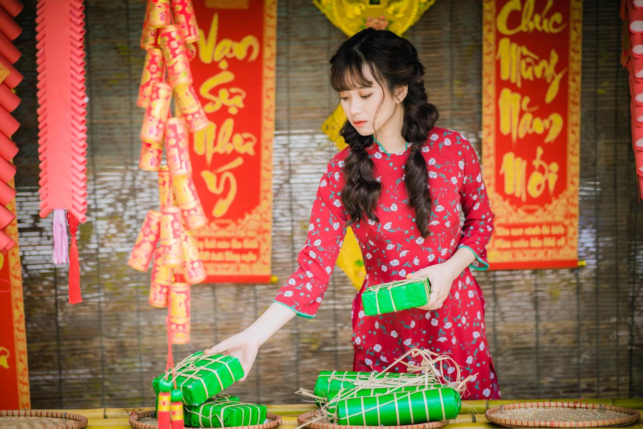 棕色双马尾卷发，齐刘海儿，穿着红色旗袍的越南少女在竹屋前，低头整理绿色的年货包 站姿 全身照 4/3侧面照 横幅 中国结 春联 鞭炮